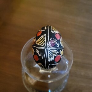 Peut inclure: Un œuf noir décoré de lignes géométriques blanches dessinées à la main, de formes triangulaires rayées jaunes et d'accents circulaires rouges. L'œuf est placé verticalement, révélant un motif symétrique divisé par des lignes blanches.