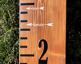 May include: A wooden growth chart with black markings and white arrows. The chart has the numbers "2" and "6 mos" written on it. The words "one" and "birth" are also written on the chart.