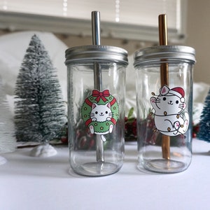 Op de afbeelding: Twee glazen potten met metalen deksels en herbruikbare rietjes. De potten hebben feestelijke kerstkat stickers. Eén kat draagt een kerstmanmuts en de andere houdt een krans vast.