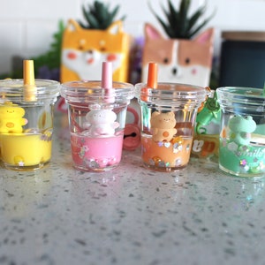 以下が含まれることがあります： 透明なプラスチック製のカップが4つあり、カラフルな液体と小さな人形が入っています。各カップにはストローとチャームが上部に付いています。チャームは、黄色のクマ、ピンクのクマ、オレンジのクマ、緑のカエルです。カップは白いカウンタートップの上に置かれています。