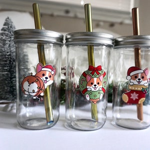 Peut inclure: Trois bocaux en verre transparent avec des couvercles en métal et des pailles réutilisables. Chaque bocal a un autocollant sur le thème de Noël différent représentant un corgi de dessin animé portant un chapeau de Père Noël.