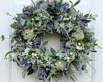 May include: A circular wreath composed of artificial foliage and flowers. The wreath incorporates sage green, lavender, and white elements. It is displayed on a white door.