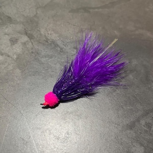 Puede incluir: Una mosca de pesca hecha a mano con un diseño vibrante. La mosca presenta un grupo de plumas de color púrpura oscuro, una cabeza rosa brillante y un pequeño anzuelo rojo. Esta mosca está diseñada para atraer peces.