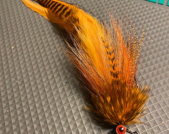 May include: A brown and orange fly fishing lure with a red bead and a black hook. The lure has a long tail of feathers and a body made of fur.
