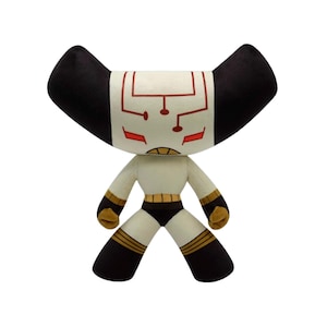Puede incluir: Un peluche con un diseño único. La figura tiene un cuerpo color crema, detalles en negro y dorado, y una cabeza distintiva con cuernos negros y una cara con detalles rojos y blancos. El juguete está sobre un fondo blanco.