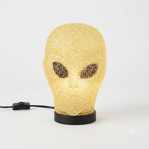 May include: A table lamp shaped like an alien head with a textured, translucent yellow surface. The lamp features two large, dark, oval-shaped eyes with a pebbled texture and sits on a circular black base with an attached black power cord.