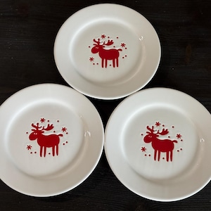 May include: Three white ceramic plates with a red moose design in the center. The moose is surrounded by small red snowflake-like shapes. The plates are arranged on a dark wooden surface.