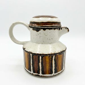 Könnte beinhalten: Kaffeekanne aus Keramik mit Deckel. Die Kanne ist weiß mit braunen Sprenkeln und einem braun-beigen gestreiften Boden. Sie hat einen gebogenen Griff und einen Ausguss.