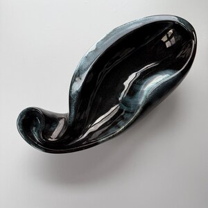 May include: A glossy, black ceramic dish with a unique, flowing shape. The dish has a deep, curved design with a subtle blue-green edge. The interior reflects light, highlighting its smooth surface. This decorative piece is ideal for serving or display.