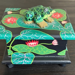 Pode incluir: Uma caixa de madeira pintada à mão com uma figura de sapo. A caixa é preta com folhas de nenúfar verdes e flores vermelhas. O sapo é verde com manchas pretas e pés bege.