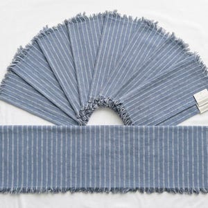 Op de afbeelding: Een set blauw-wit gestreepte stoffen servetten en een bijpassende tafelloper. Elk item heeft een verticaal krijtstreepmotief en gerafelde randen aan de uiteinden. De artikelen zijn in een waaier gerangschikt met een productlabel bevestigd.