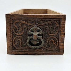 Könnte beinhalten: Eine rechteckige Holzschublade mit dunkelbrauner Oberfläche und kunstvollen Schnitzereien in Form von Rankenmustern auf der Vorderseite. Ein mittiger Metall-Fallgriff mit verwitterter Patina ist an der geschnitzten Holzfront befestigt.