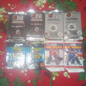 Könnte beinhalten: Eine Sammlung ungeöffneter Hockey-Sammelkartenpackungen. Die Packungen stammen von verschiedenen Marken und Jahren, darunter Pro Set, Score und Stadium Club. Die Packungen liegen auf einer roten und grünen Tischdecke mit Blumenmuster.