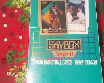 May include: A teal blue box with a Skybox Series II logo and the text "NBA Basketball Cards 1990-91 Season". The box features a basketball card of Akeem Olajuwon, a player for the Houston Rockets, wearing a red and white jersey with the number 31.