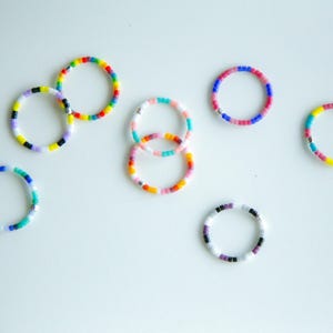 Könnte beinhalten: Eine Sammlung bunter Perlenringe in verschiedenen Größen und Mustern. Die Ringe weisen eine Vielzahl von Farben auf, darunter Blau, Grün, Gelb, Rot, Rosa und Lila. Die Ringe sind auf einer weißen Oberfläche angeordnet.