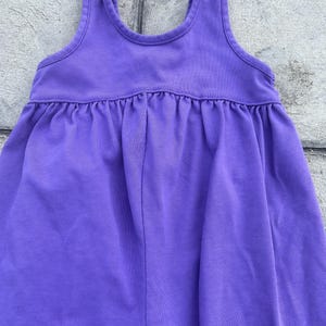 以下が含まれることがあります： ノースリーブの紫色のドレス。丸いネックラインとギャザーウエストが特徴です。柔らかく無地の生地で作られています。スカート部分はわずかにフレアになっています。