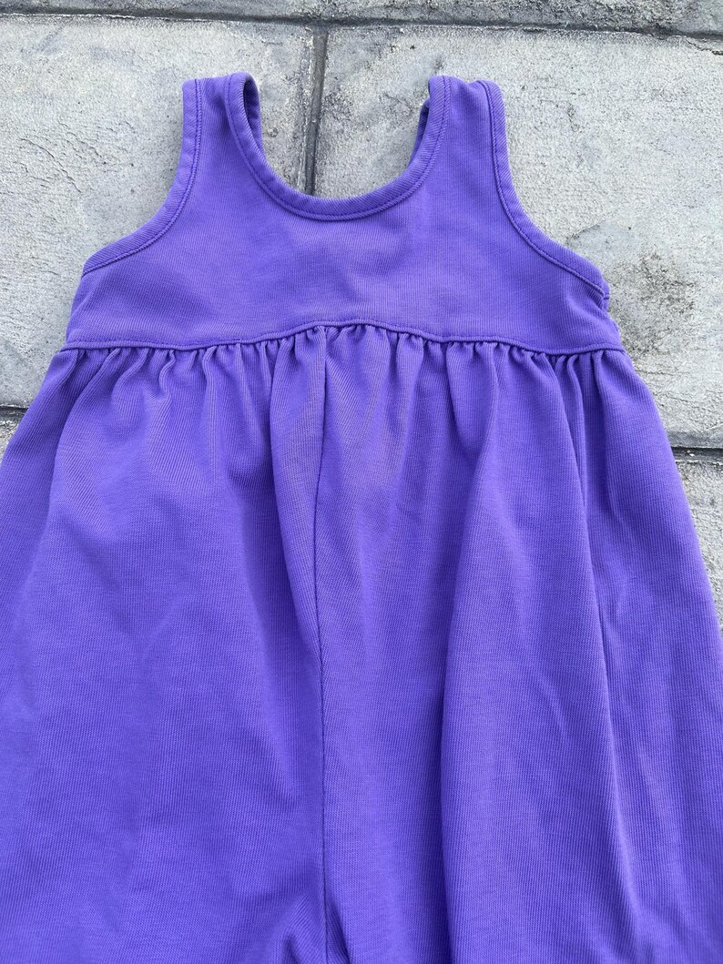 以下が含まれることがあります： ノースリーブの紫色のドレス。丸いネックラインとギャザーウエストが特徴です。柔らかく無地の生地で作られています。スカート部分はわずかにフレアになっています。