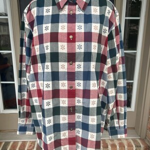 May include: A long-sleeved button-down shirt featuring a multi-colored plaid pattern in shades of dark green, deep red, blue, and white. The fabric is decorated with a repeating small white flower motif centered within each plaid square.