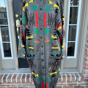 May include: A long-sleeved, knee-length knit cardigan in charcoal gray featuring a vibrant geometric pattern in red, yellow, and green. The garment includes a full-length front button closure, a round neckline, and two large patch pockets.