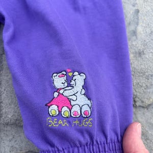 以下が含まれることがあります： 紫色の袖のアップ。刺繍のデザインは、ピンクのドレスを着たテディベアと白いテディベアが抱き合っている様子で、上にはハートがあります。「BEAR HUGS」の文字が刺繍されています。