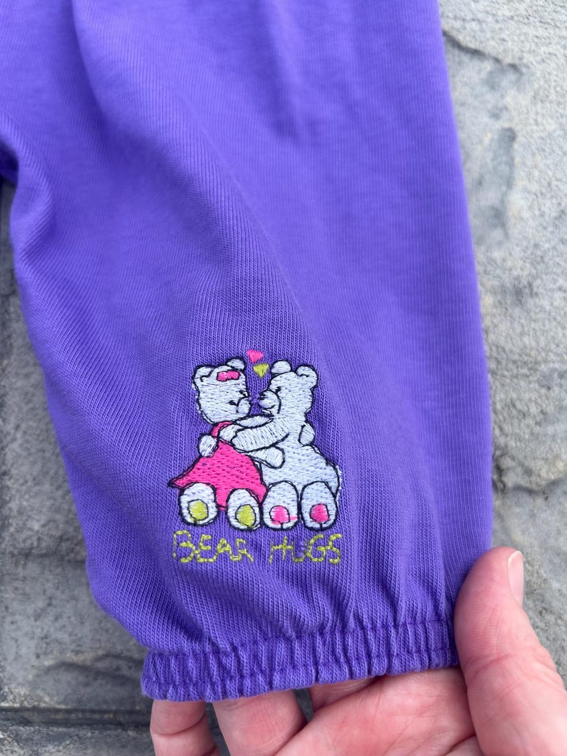 以下が含まれることがあります： 紫色の袖のアップ。刺繍のデザインは、ピンクのドレスを着たテディベアと白いテディベアが抱き合っている様子で、上にはハートがあります。「BEAR HUGS」の文字が刺繍されています。