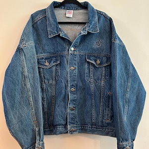 May include: A vintage denim jacket in a medium wash blue. The jacket features a button-up front, two chest pockets, and two side pockets. A small logo is embroidered on the upper left chest. The jacket is hanging on a black hanger.