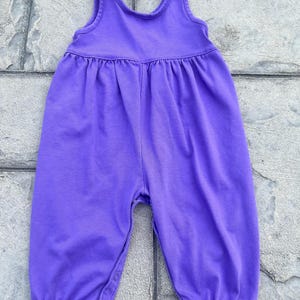 以下が含まれることがあります： 袖なしデザインの無地の紫色のベビーロンパース。 ロンパースは、ギャザーウエストラインと股下のスナップで、おむつ替えが簡単です。 脚の開口部とネックラインはシンプルな裾で仕上げられています。
