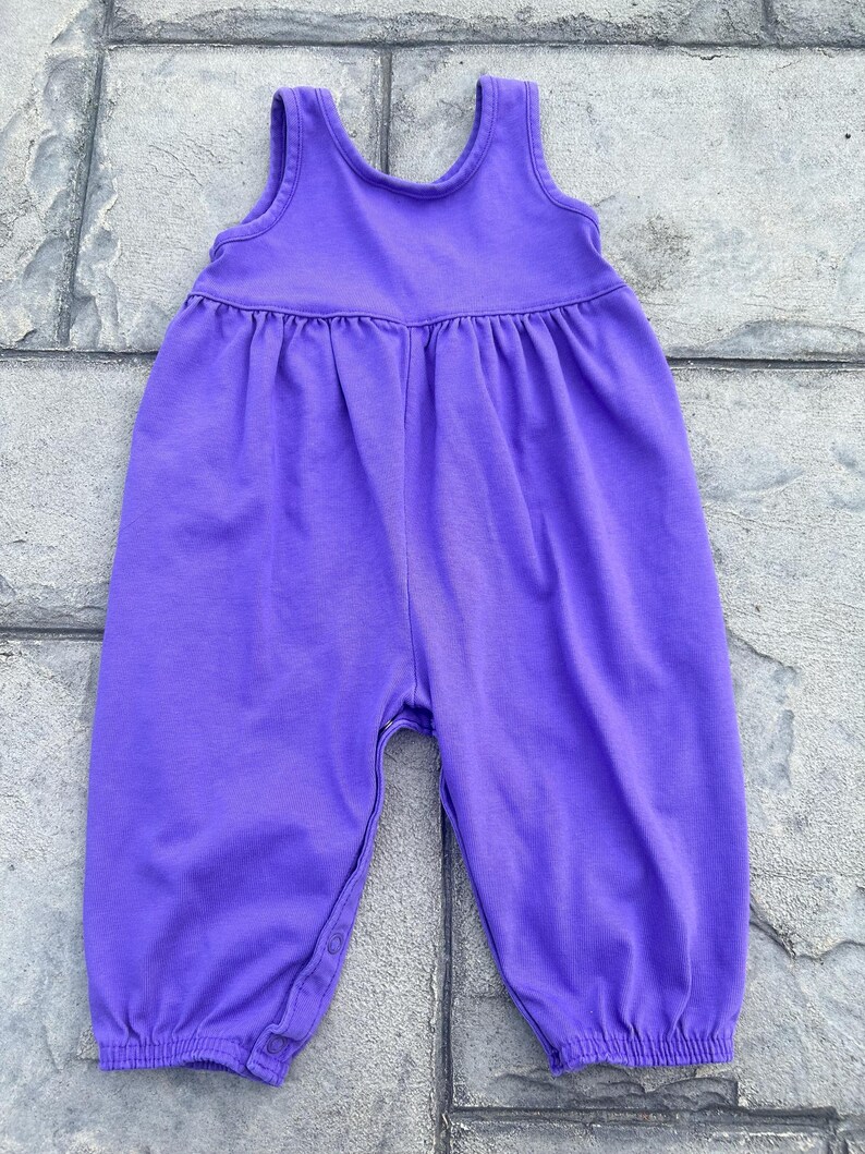 以下が含まれることがあります： 袖なしデザインの無地の紫色のベビーロンパース。 ロンパースは、ギャザーウエストラインと股下のスナップで、おむつ替えが簡単です。 脚の開口部とネックラインはシンプルな裾で仕上げられています。