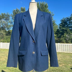 May include: A navy blue blazer with two front pockets and a two-button closure. The blazer is displayed on a mannequin outdoors against a backdrop of a blue sky and green trees. The blazer has a classic, tailored design.