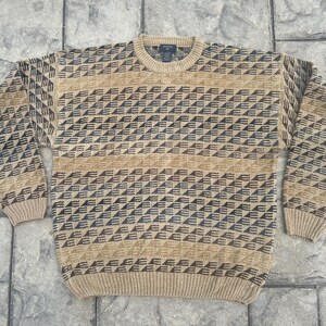 May include: A long-sleeve crew neck sweater featuring a repeating geometric pattern of dark brown and tan triangles. The knit garment has a ribbed collar, cuffs, and hem, with a tan base color and a consistent horizontal band design throughout.
