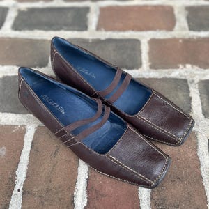 May include: A pair of dark brown leather flats with a square toe and two thin elastic straps across the top. The interior lining is blue and features the Aerosoles brand name printed in white. The shoes have visible white contrast stitching.