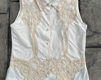 May include: A sleeveless white blouse with intricate beige lace detailing. The blouse features a collared neckline, pearl buttons, and delicate lace patterns on the front. The label reads "E. Clair Designs".