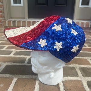 May include: A baseball cap covered in red, white, and blue sequins. The brim features horizontal stripes in white and red, while the crown is blue with white sequined stars scattered across the surface, mimicking a national flag pattern.
