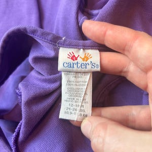 以下が含まれることがあります： 紫色のベビー服のクローズアップ。タグには、Carter'sのロゴと手形、英語とスペイン語のテキストが記載されています。タグには、衣類が綿100％で、12〜18ヶ月用で、10.9〜11.8kgであることが示されています。