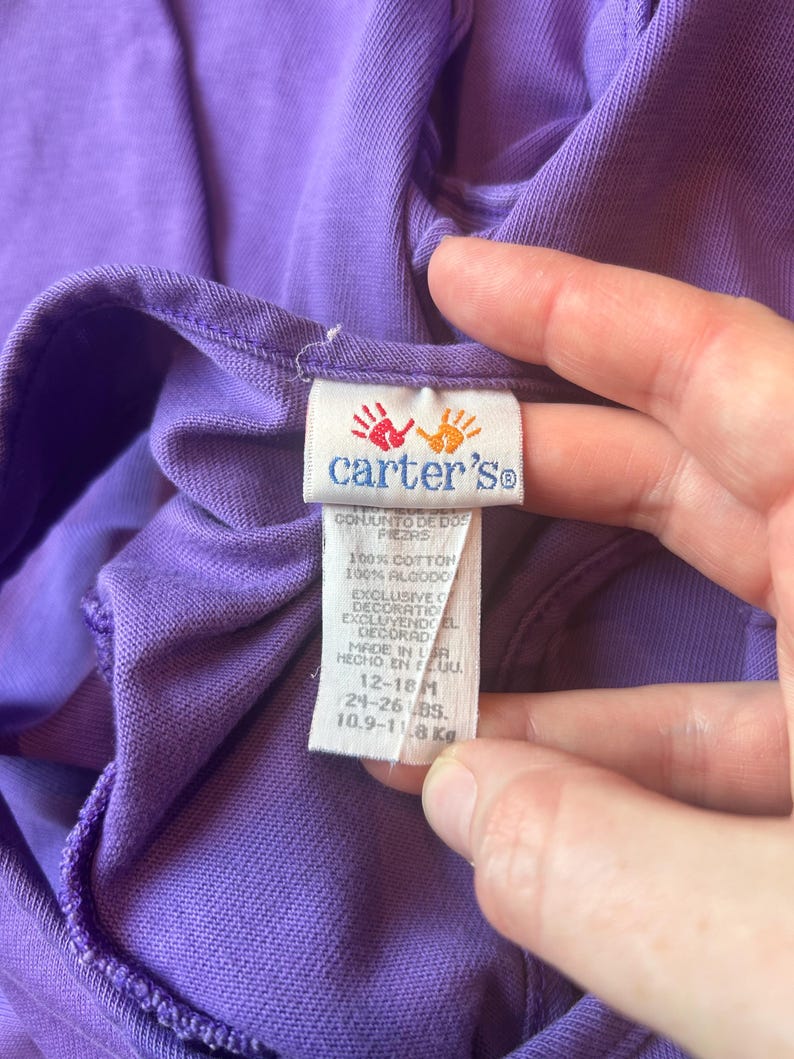 以下が含まれることがあります： 紫色のベビー服のクローズアップ。タグには、Carter'sのロゴと手形、英語とスペイン語のテキストが記載されています。タグには、衣類が綿100％で、12〜18ヶ月用で、10.9〜11.8kgであることが示されています。