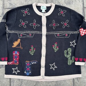 May include: Black button-up cardigan sweater with embroidered Southwestern designs. Features cacti, stars, hearts, and cowboy boots in red, white, and tan. The sweater has a cream-colored trim.