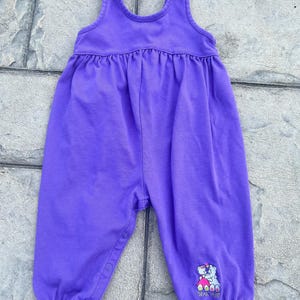 以下が含まれることがあります： 袖なしの紫色のベビー用ジャンプスーツ。ウエストにギャザー、脚にスナップ留めが付いています。右下の脚には、ハートを持った2匹のクマの小さな刺繍デザインがあります。生地は柔らかいニット素材のようです。
