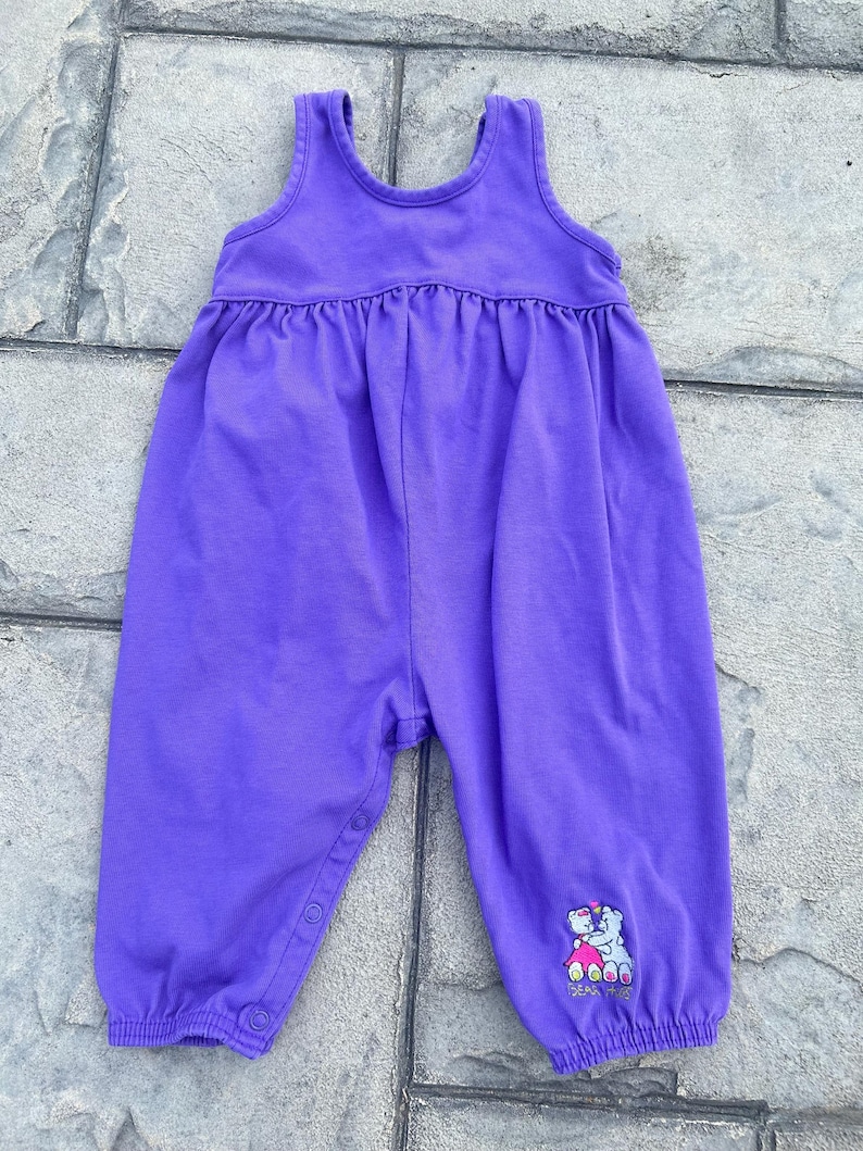 以下が含まれることがあります： 袖なしの紫色のベビー用ジャンプスーツ。ウエストにギャザー、脚にスナップ留めが付いています。右下の脚には、ハートを持った2匹のクマの小さな刺繍デザインがあります。生地は柔らかいニット素材のようです。