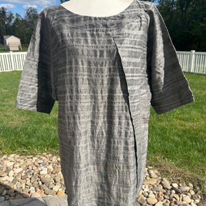 May include: A gray, short-sleeved tunic with a unique asymmetrical design. The fabric has a subtle striped texture. The tunic is displayed on a mannequin outdoors, showcasing its drape and style. The tunic's color is a neutral gray.