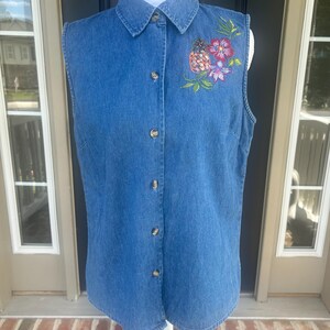 May include: A sleeveless blue denim button-down shirt featuring a pointed collar and a front button closure. The upper left chest area is decorated with an embroidered design of a pineapple and colorful flowers in shades of pink and purple.