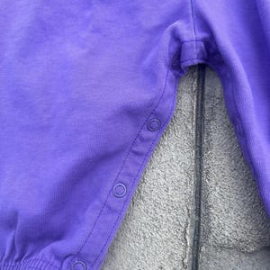 以下が含まれることがあります： 紫色のベビーロンパースのクローズアップ。 ロンパースは柔らかい生地で作られており、着脱しやすいように側面にスナップ留めが付いています。 この衣服は、ギャザーの入った伸縮性のあるウエストバンドと脚の開口部を備えています。