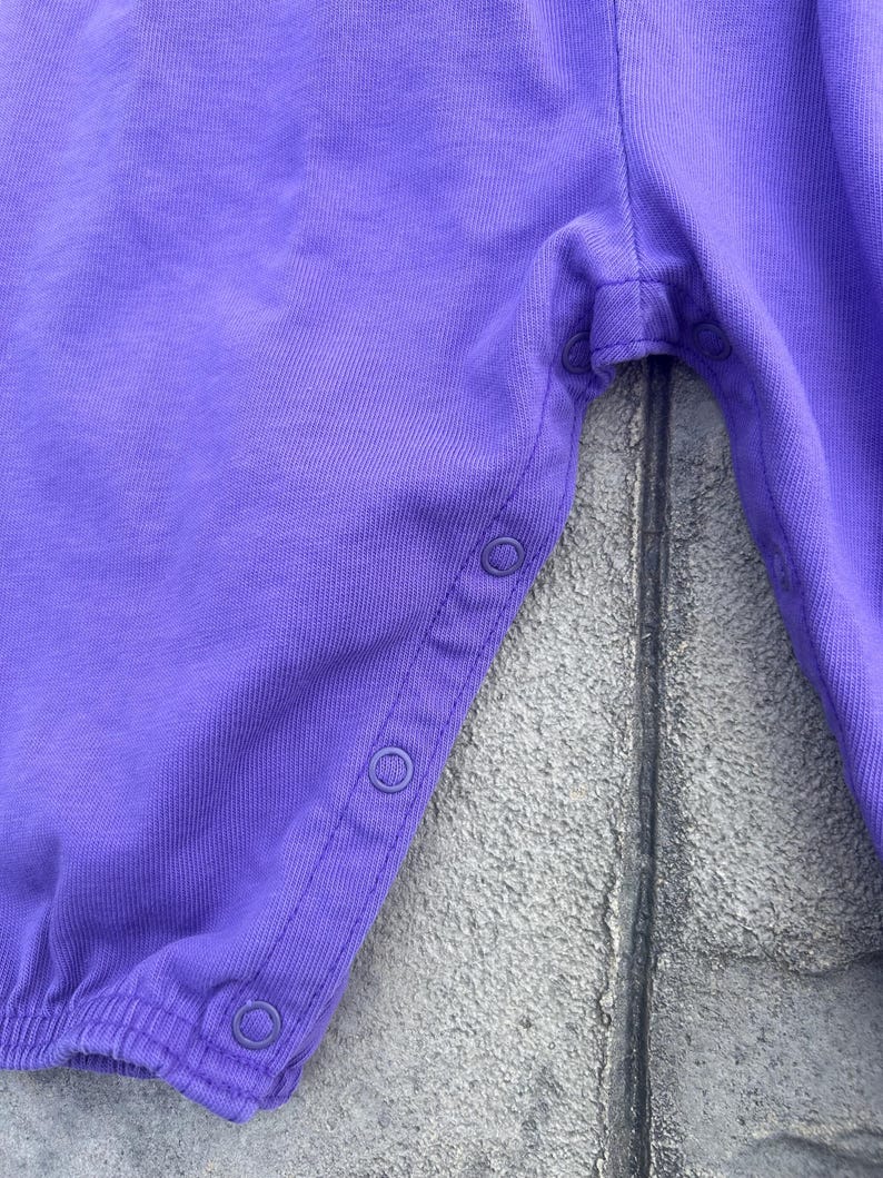 以下が含まれることがあります： 紫色のベビーロンパースのクローズアップ。 ロンパースは柔らかい生地で作られており、着脱しやすいように側面にスナップ留めが付いています。 この衣服は、ギャザーの入った伸縮性のあるウエストバンドと脚の開口部を備えています。