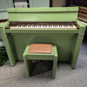 May include: An upright piano finished in a matte sage green colour with a matching bench featuring a brown leather-textured seat. The piano has a hinged lid, a keyboard with white and black keys, and silver-toned metal accents on the side pillars.