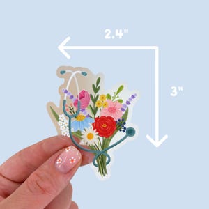 Może przedstawiać: Przezroczysta naklejka przedstawiająca bukiet kolorowych kwiat&oacute;w i turkusowy stetoskop. Naklejka ma wymiary 6,1 cm x 7,6 cm. Kwiaty obejmują czerwone r&oacute;że, r&oacute;żowe i niebieskie stokrotki oraz gałązki lawendy. Naklejka jest trzymana przez dłoń.