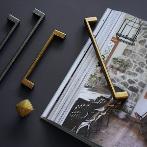 May include: Four cabinet hardware pulls in two different finishes. Two gold pulls are rectangular with rounded corners. Two black pulls are rectangular with squared corners. One gold knob is square with a diamond pattern.