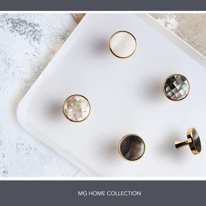 May include: Five decorative cabinet knobs with a gold finish. The knobs are made of mother of pearl and have a variety of patterns.  MG HOME COLLECTION is printed below the knobs.