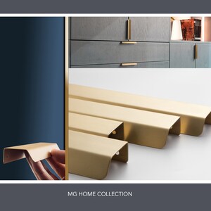 May include: A set of five gold-toned cabinet pulls with a curved edge. The pulls are arranged in a row on a white surface. The text "MG HOME COLLECTION" is printed below the pulls.