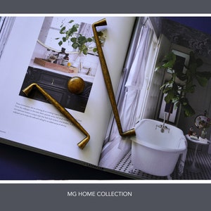May include: Two gold metal cabinet door pulls with a curved design. The pulls are shown on a page from a home decor magazine. The magazine page shows a bathroom with a white bathtub and a large plant.