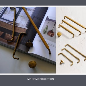 May include: A collection of antique gold cabinet hardware, including pulls and knobs. The hardware is displayed on a white surface and a book with a rustic interior design photo. The text "MG HOME COLLECTION" is printed below the image.