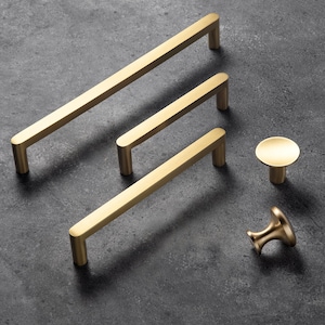 May include: A selection of gold-coloured cabinet hardware, featuring three rectangular bar pulls of different lengths and two knob styles. The hardware is displayed on a dark grey surface, showcasing a modern design aesthetic.